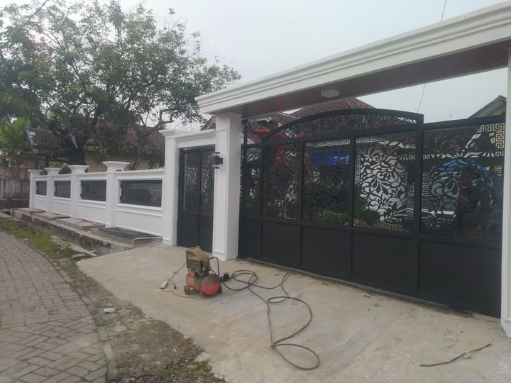 Pagar besi hitam bermotif ukir dengan tembok putih di sebuah rumah